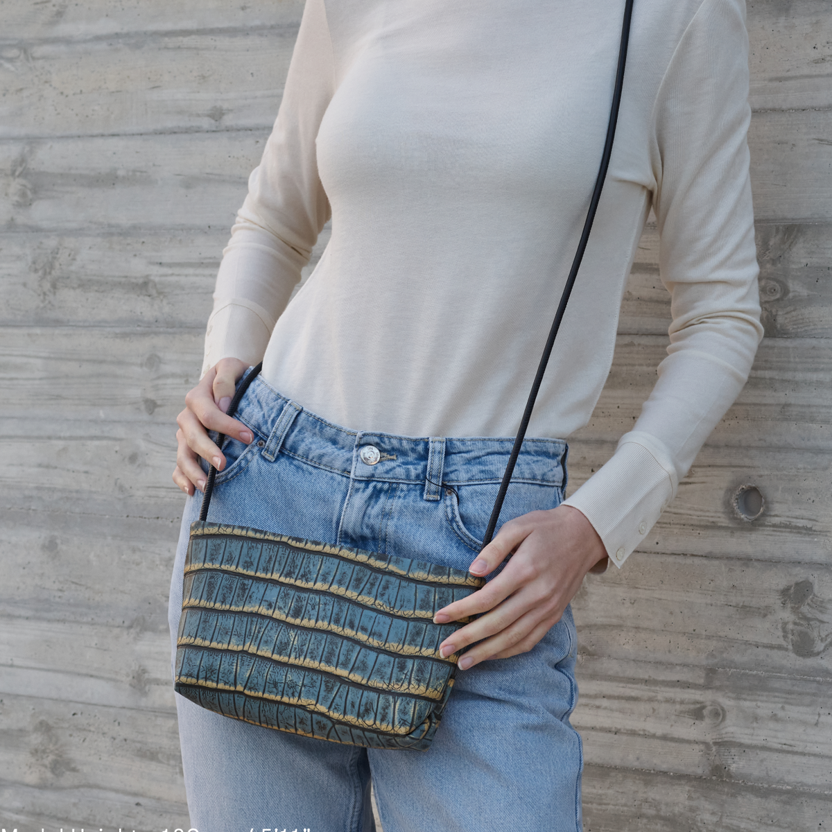 אישה עם תיק צד כחול קטן בדוגמת קרוקודיל על רקע קיר בטון.